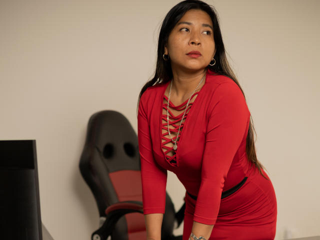 LunnaEvans with long black hair, red dress, lace-up neckline, and hoop earrings, leaning forward, looking to the side, showing her tits and a slight curve to her ass.