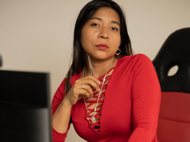 LunnaEvans with medium tits, wearing a red lace-up top, holding a pen, looking slightly bored, sitting in a red chair, with long black hair and gold hoop earrings.