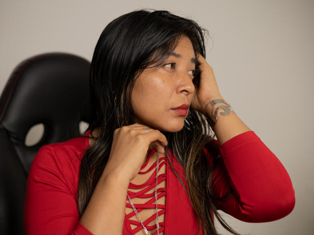 LunnaEvans with long black hair, wearing a red lace-up top, has her hand on her head, showing a tattoo on her right wrist, looking to the side.