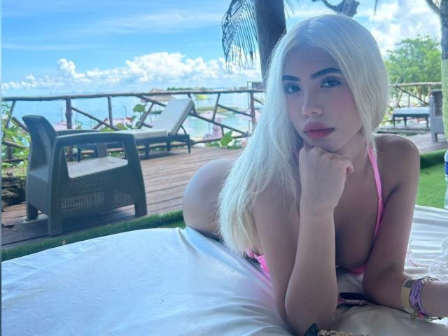 ChristieGarci with long blonde hair, pink bikini top, lying on white bed, looking at camera, hand on chin, small pink lips, blue eyes, wooden deck, clear sky, ocean.