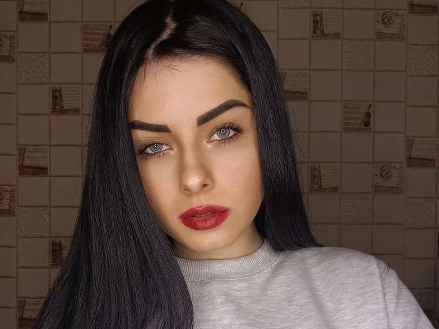 SelinaT has long black hair, thick eyebrows, green eyes, red lipstick, and a gray shirt. Her face is centered, looking slightly to the left with a serious expression.