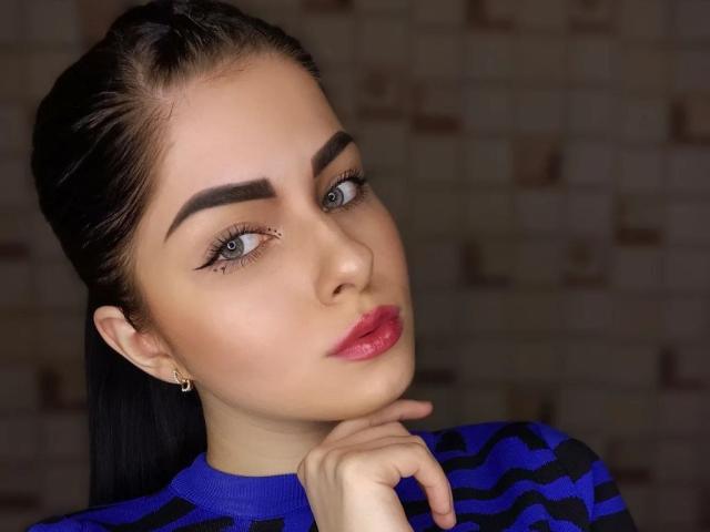 SelinaT with dark hair, thick eyebrows, green eyes, and pink lipstick, wearing a blue and black striped shirt, resting her chin on her hand.