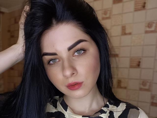 SelinaT has black hair, green eyes, and is looking at the camera with a slight pout. She's touching her hair with her right hand, wearing a camo shirt.