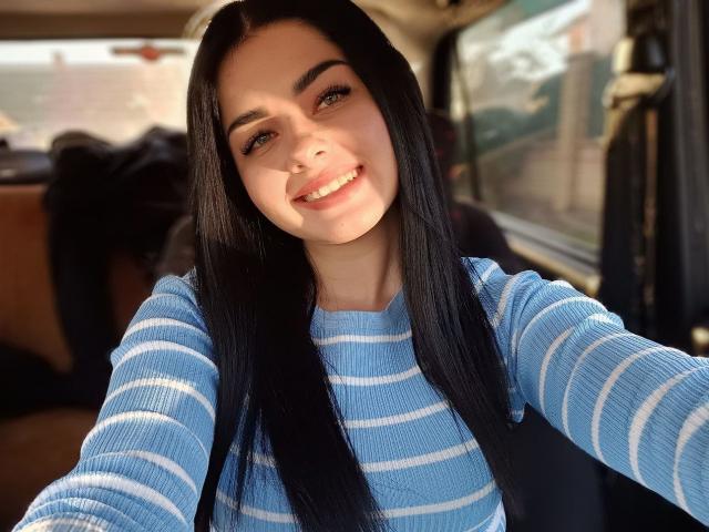 SelinaT with long black hair, blue striped shirt, smiling, light skin, green eyes, sitting in a car, taking a selfie, showing her cute face and small tits.