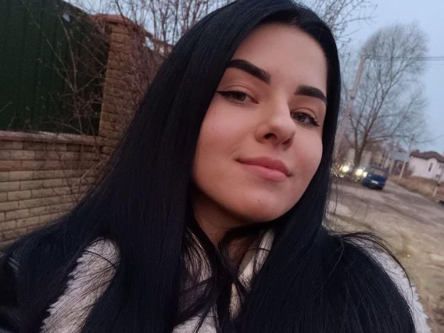 SelinaT has long black hair, brown eyebrows, and a subtle smile. She's wearing a white and black striped top. Her nose is small and she has light skin.
