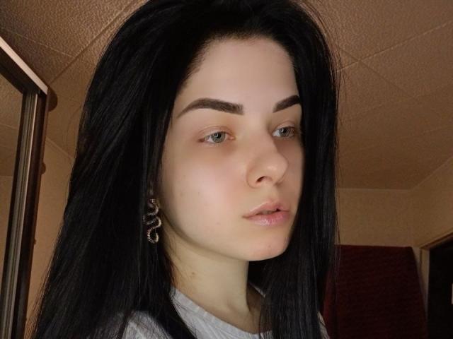 SelinaT has pale skin, black hair, green eyes, and full pink lips. She's wearing a silver earring with a heart-shaped design on her right ear.