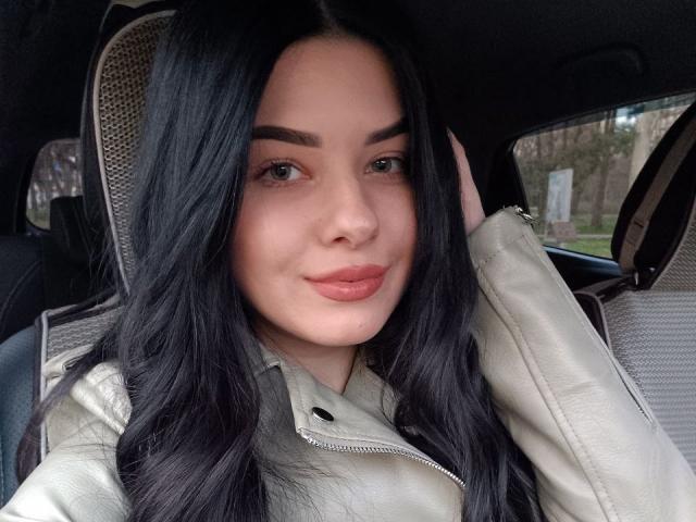 SelinaT has black hair, green eyes, and full pink lips, wearing a beige leather jacket, sitting in a car with a patterned headrest, touching her hair.
