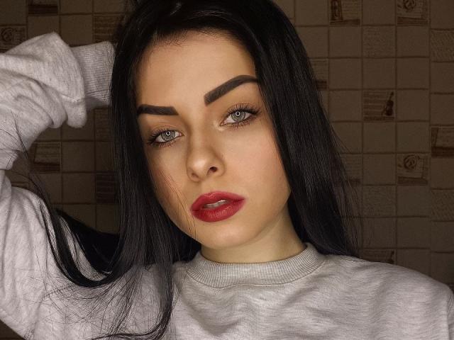SelinaT with dark eyebrows, long black hair, blue eyes, red lips, and a gray sweatshirt, looking at the camera with a serious expression, hand in hair.