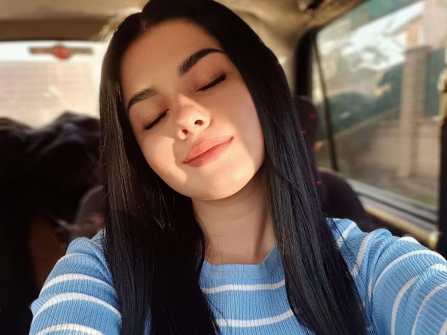 SelinaT with long black hair, eyes closed, wearing a blue striped shirt, has a slight smile, sunlit face, and a relaxed expression. Her nose is slightly visible.