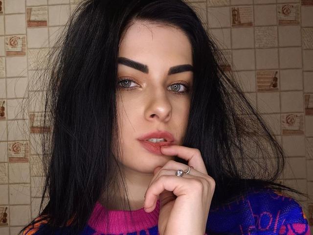 SelinaT has dark, messy hair, full eyebrows, light green eyes, and pink lips with a finger near her mouth. She's wearing a purple and blue sweater, showing a ring.