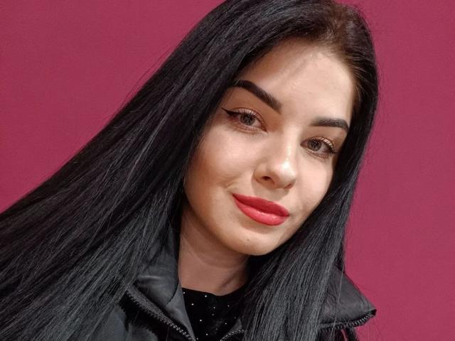 SelinaT has long black hair, thick eyebrows, red lipstick, and is wearing a black jacket over a black top with small white dots. Her eyes are slightly upturned.