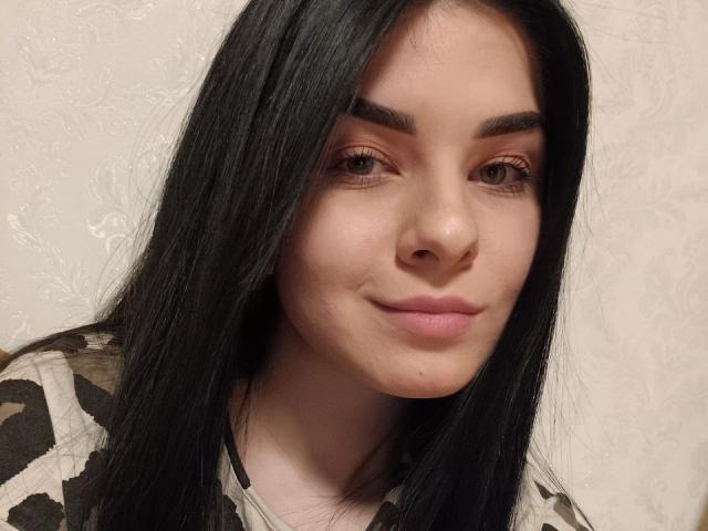 SelinaT has long black hair, brown eyes, and a slight smile. She's wearing a camouflage top with a pale, textured wall in the background.