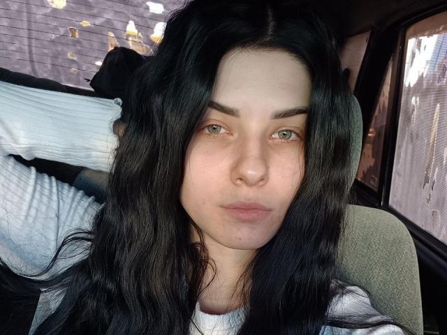 SelinaT has long black hair, green eyes, and a pouty face. She's wearing a white ribbed shirt, sitting in a car with her arm raised. Her ass is not visible.