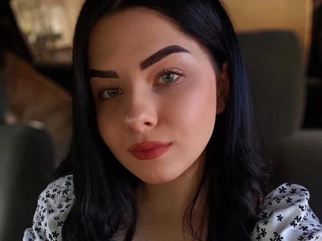 SelinaT has dark, wavy hair, thick, arched eyebrows, green eyes, and red lips. She's wearing a white, floral-patterned top, looking serious with a slight pout.