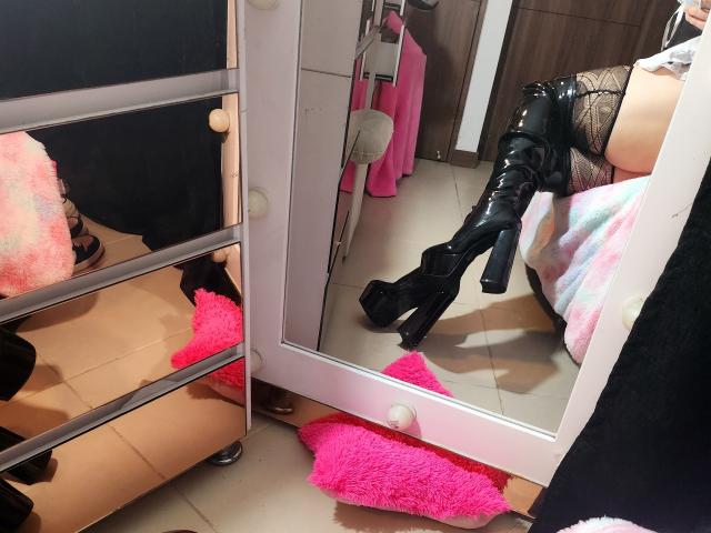 Akkanees glänzende, schwarze, hohe Stiefel mit hohen Absätzen spiegeln sich in einem Spiegel wider, sie steht auf einem rosa, flauschigen Teppich, trägt Spitzenstrümpfe und ein teilweise sichtbares, rosa Kleid..