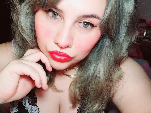 Akkanee, met groene ogen, een roze blos en rode lippenstift, heeft grijsgroen haar, een zwarte kanten top en een witte choker, en ze houdt haar kin vast met een schattige, sexy uitdrukking..