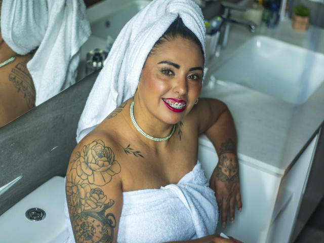 Genexis mit einem Handtuch im Haar, sitzend in einer Badewanne, lächelnd mit rotem Lippenstift, mit einer Perlenkette, die Tattoos an Schulter und Arm zeigt, mit heller Haut..