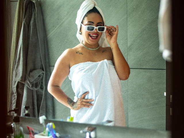 Genexis with a white towel wrapped around her ass, wearing white sunglasses, smiling, hand on hip, tattoo on her chest, towel on her head, standing in a bathroom.
