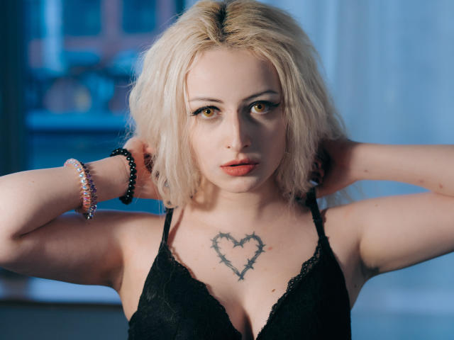 NateLove with blonde hair, black lace top, heart tattoo on chest, arms raised, wearing bracelets, staring into camera with serious expression, light makeup, blue eyes, slightly parted lips.