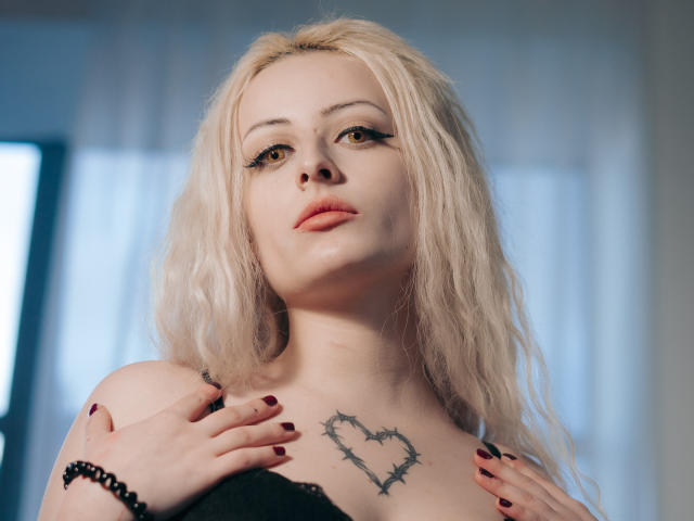 NateLove has pale skin, blonde wavy hair, and a heart tattoo with barbed wire above her tits. She's looking up with dark eyeliner, red lipstick, and maroon nails.