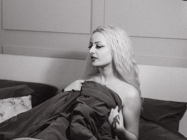 NateLove with long blonde hair, bare shoulders, and a tattoo on her right hand, holding a dark blanket over her tits, sitting on a bed, looking to the side.