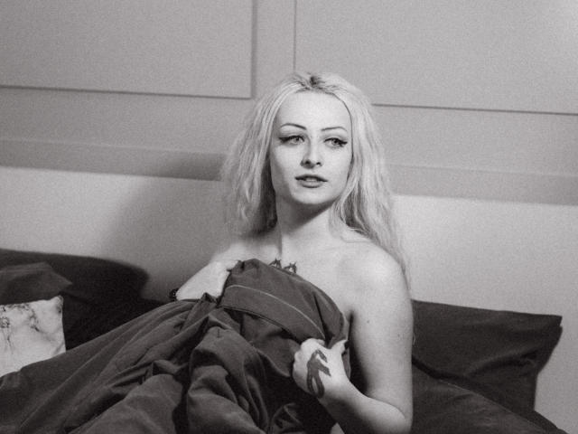 NateLove has pale skin, long blonde hair, and is nude with a blanket covering her tits, sitting in bed, holding the blanket with a sexy, slightly open-mouthed look.