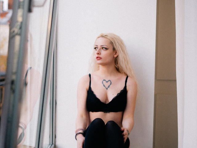 NateLove with blonde hair, black lace bra, heart tattoo on chest, and black leggings, sitting against a white wall, looking to the side with a slightly pouty expression.
