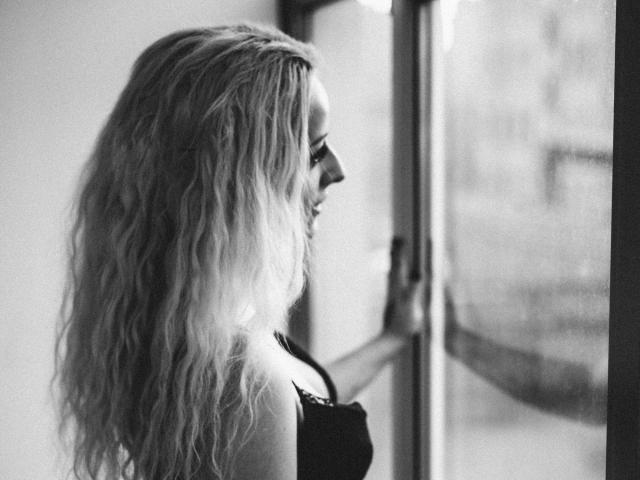 Black-and-white photo of NateLove with long, wavy blonde hair, looking out a window, wearing a black top, showing her small tits and smiling softly.