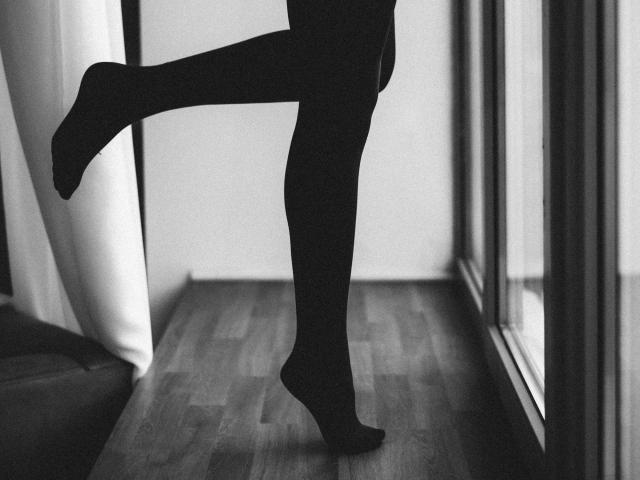 NateLove's black panty-clad legs are lifted, showing a raised heel, standing on wooden floor, near a window, in a black-and-white photo.