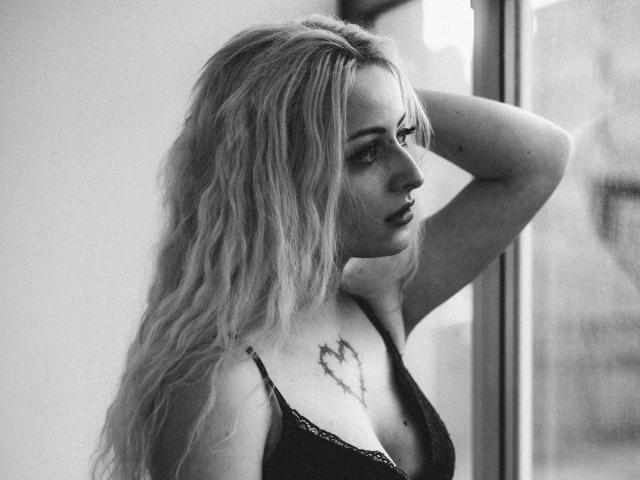 Black-and-white photo of NateLove with wavy blonde hair, dark lipstick, tattoo on chest, hand on head, looking out window, wearing a black lace bra, showing small tits.