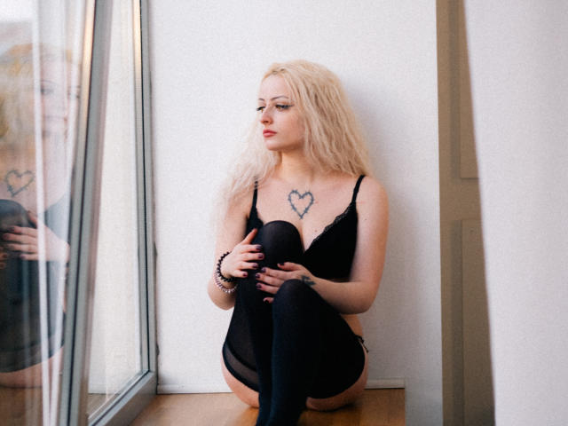 NateLove with blonde hair, black lingerie, and thigh-high socks, sitting on window ledge, holding knees, heart tattoo on chest, looking to the side, reflection in glass.