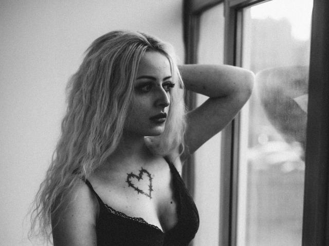 NateLove with wavy blonde hair, black lace bra, heart tattoo on chest, arm raised, looking out window, black and white, sexy, serious expression.