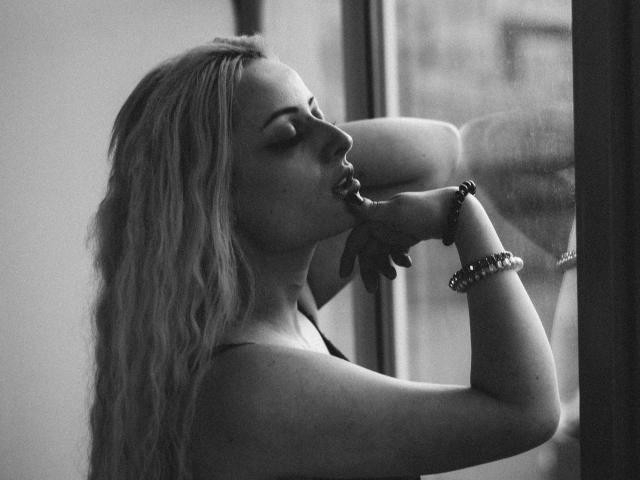 Black-and-white photo of NateLove with long, wavy blonde hair, pouty lips, and eyes closed, sucking her finger, wearing a bracelet, standing by a window, in profile.