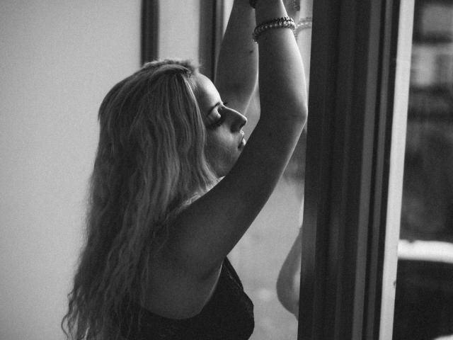 NateLove with wavy blonde hair, arms raised, looking out a window, wearing a black lace top, bracelet on wrist, in a black-and-white shot.