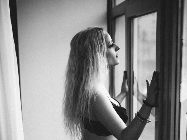 Black and white photo of NateLove with wavy blonde hair, wearing a lacy bra, standing by a window, touching glass, looking outside, wrist bracelet, side profile, nipples visible.