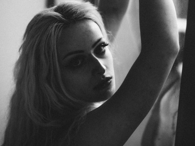 Black-and-white photo of NateLove with long, blonde hair, arms raised, looking seductively at the camera, showing pouty lips and slightly parted, dark, sultry eyes.