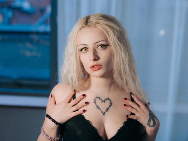 NateLove with blonde hair, black lace bra, and thorny heart tattoo on chest, hands on chest, red nails, bracelet, looking at camera, soft blue background.