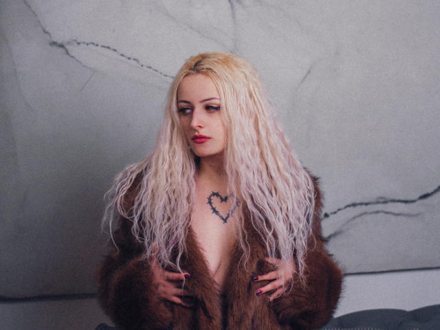 NateLove with long, wavy blonde hair, wearing a brown fur coat, exposing her tits, has a heart tattoo on her chest, and red lipstick, looking pouty.