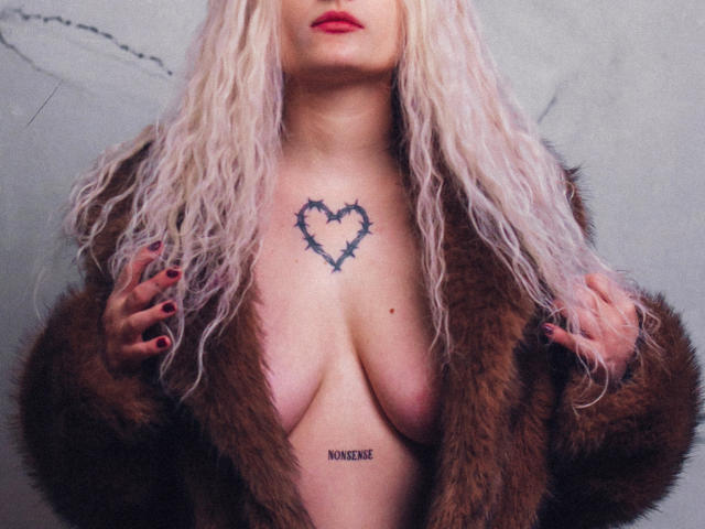 NateLove with blonde wavy hair, red lipstick, brown fur coat open showing tits, thorny heart tattoo, mons tattoo, hands in coat, red nails.