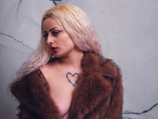 NateLove with blonde hair, wearing a brown fur coat, exposing her tits, heart tattoo on chest, red lipstick, and eyeliner, looking down, against a gray wall.
