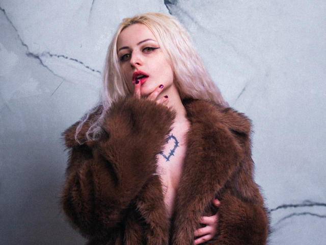 NateLove with blonde hair, wearing a brown fur coat, biting her finger, showing a heart tattoo on her chest, red lipstick, and dark nail polish.