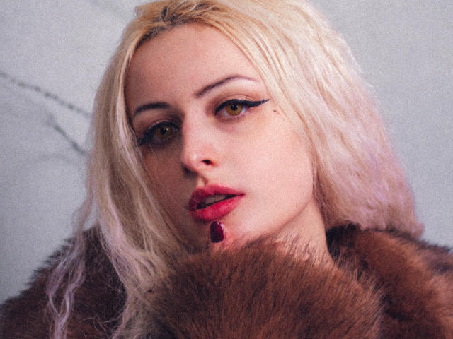 NateLove with blonde hair, red lipstick, and black eyeliner, wearing a brown fur coat, has a red jewel earring, and a sultry look in a grainy photograph.