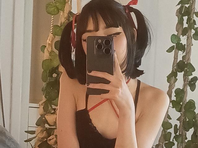 SilencePsi with puffy black twin-tails, red ribbons, black lace top, holding phone, covering face, showing small tits, taking selfie, green vines in background, holding phone with three cameras.