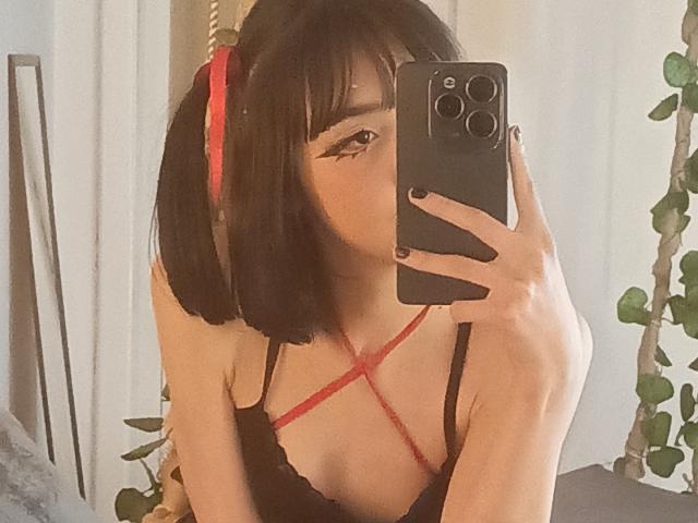 SilencePhsi with black bob, red ribbon in hair, black lace bra, criss-cross red straps, taking selfie in mirror, holding black phone, cute, slender, fair skin, subtle eyeliner.