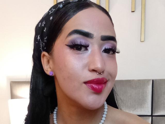MichellDiaz ha labbra rosa lucide, sopracciglia nere folte, ombretto viola, ciglia lunghe, capelli neri con un foulard a motivi stellati e un piercing al naso, e indossa una collana di perle..