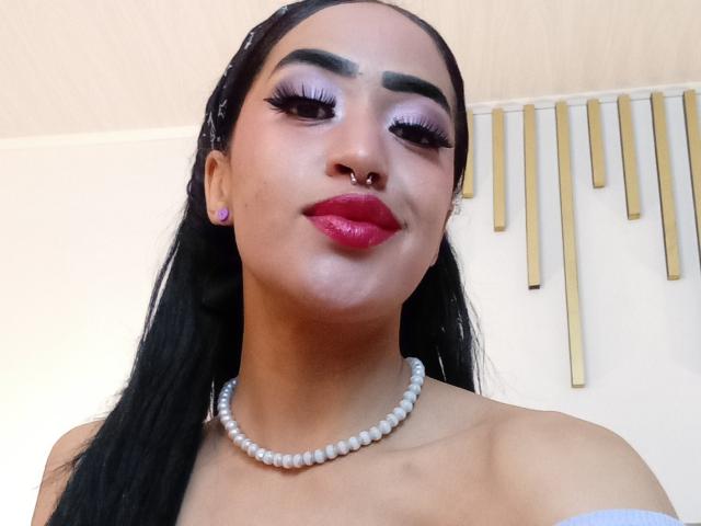 MichellDiaz tem cabelo escuro, pálpebras roxas, cílios longos, batom vermelho, um piercing no nariz, brincos rosa, um colar de pérolas brancas e um sorriso confiante. Sua pele é de cor clara..
