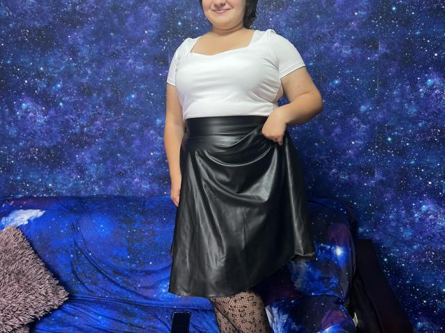 AlisaMi with medium tits, white shirt, black leather skirt, holding it up, standing, smiling, wearing patterned tights, in front of a blue starry wall and blue sofa.