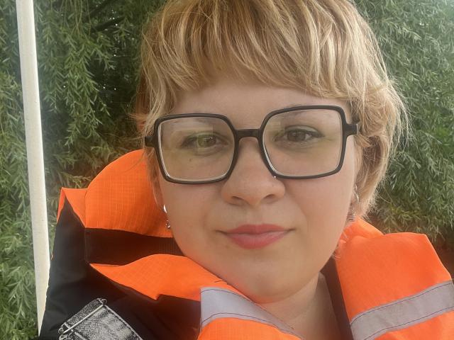 AlisaMi with short blonde hair, wearing black glasses and an orange safety vest, has a slight smile and pink lipstick, her face is close to the camera.