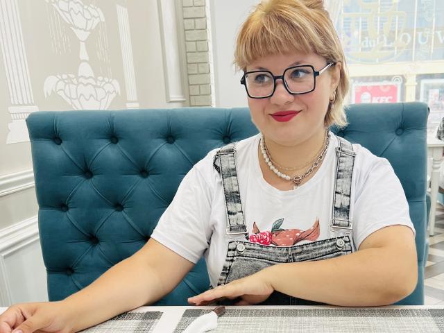 AlisaMi with short blonde hair, glasses, red lipstick, wearing a white shirt with a crab graphic and denim overalls, sitting in a blue tufted chair, smiling.