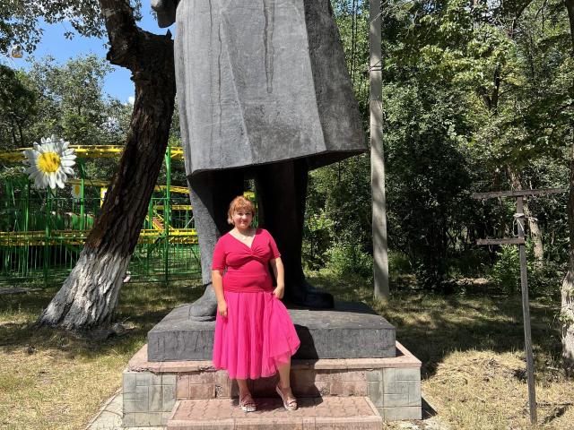 AlisaMi stands in front of a tall black statue with a wide base, wearing a pink dress and red shoes, her short red hair visible, in a sunny park.